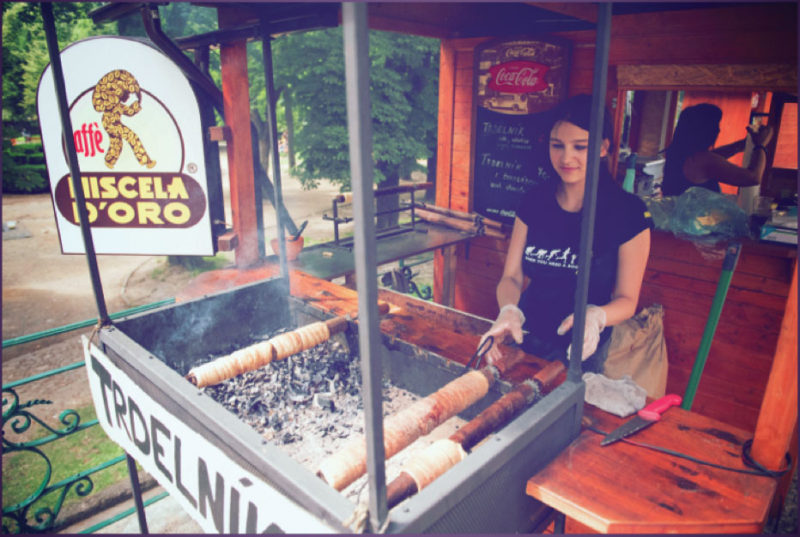 A young local prepares the popular snack, tredlnik, wrapping strings of dough on a wood stick and cooking on hot coals. The freshly baked pastry is rolled in sugar and ground nuts